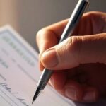 Une main qui remplit un chèque avec un stylo (Crédit : Alex.I)